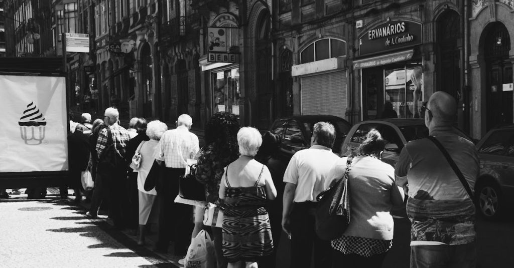 Monochrome street photo capturing a queue of people standing in front of old buildings in an urban setting.